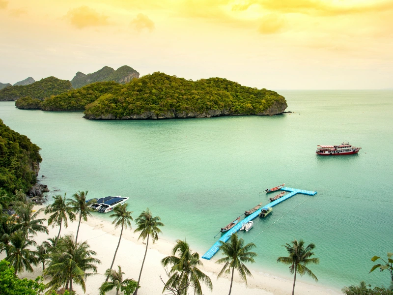 Scenic tropical beach with turquoise water, palm trees, small islands in the background, and a floating dock with boats during a golden sunrise or sunset.