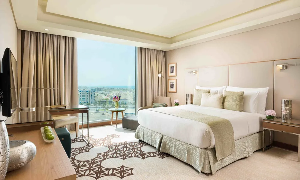 Luxurious guest room at Grand Millennium Muscat featuring a king-size bed with elegant beige linens, modern decor, large window with city views, and stylish furnishings
