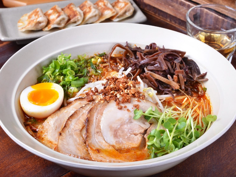 at Tokyo bowl of ramen topped with sliced pork, soft-boiled egg halves, green onions, bean sprouts, mushrooms, and microgreens, with a side of gyoza and a glass of tea in the background - Japan Travel 2026