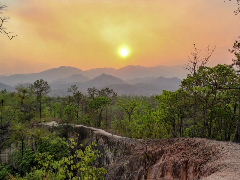 Sunset over a mountain range with warm orange and yellow hues, viewed from a cliff edge surrounded by lush green trees.