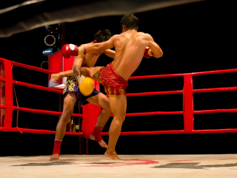 Two Muay Thai fighters in a boxing ring, one in red shorts delivering a high kick to the opponent in blue and yellow shorts, against a dark background.