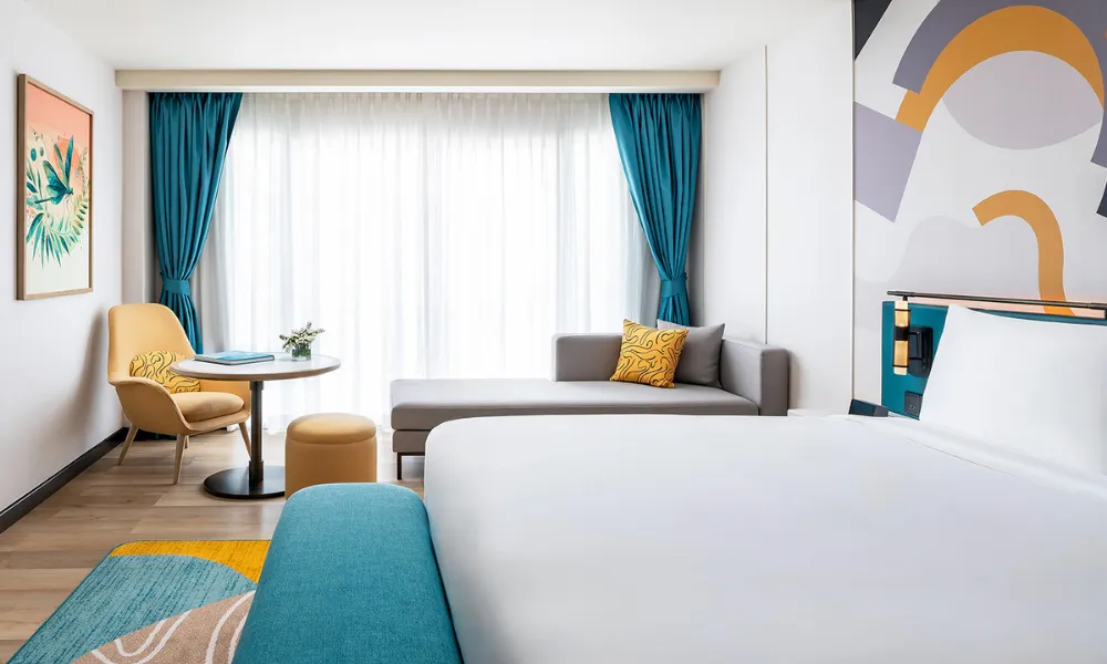 Modern hotel room at M Social Phuket featuring a king-size bed, vibrant abstract wall art, teal and mustard accents, a cozy lounge chair with a round table, and a chaise by a large window with sheer curtains.