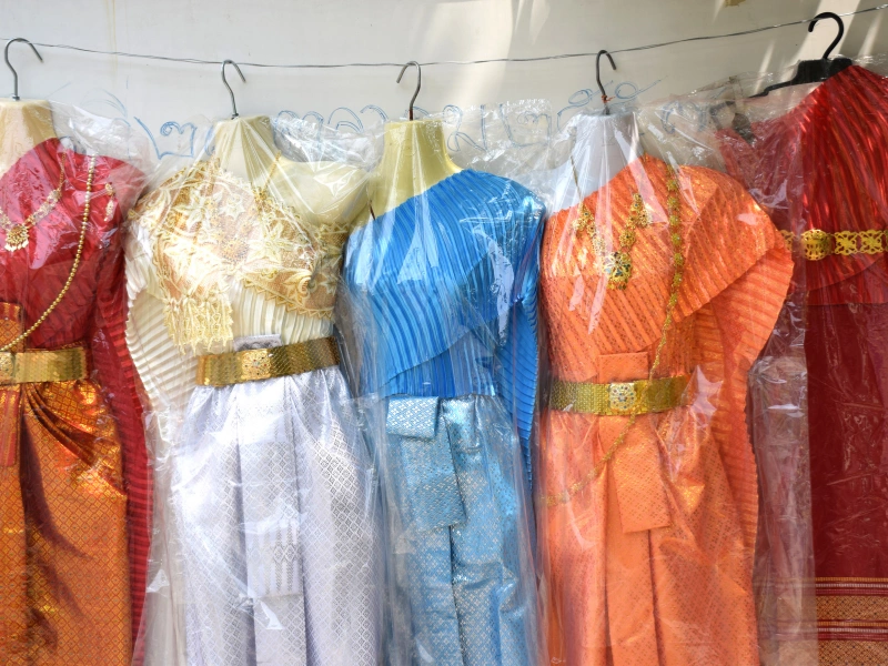 The image shows five traditional Thai dresses hanging on mannequins. Each dress is covered in a clear plastic garment bag. The dresses are in various colors: red, gold, blue, orange, and another red dress with different patterns and designs. They all have intricate details and are accessorized with golden belts.