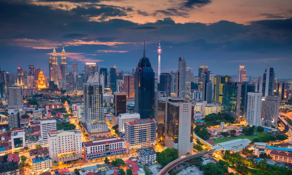 A panoramic view of Kuala Lumpur's skyline at dusk, showcasing a mix of modern skyscrapers and historic buildings. The iconic Petronas Twin Towers and the Kuala Lumpur Tower are prominently visible against a backdrop of a colorful evening sky.