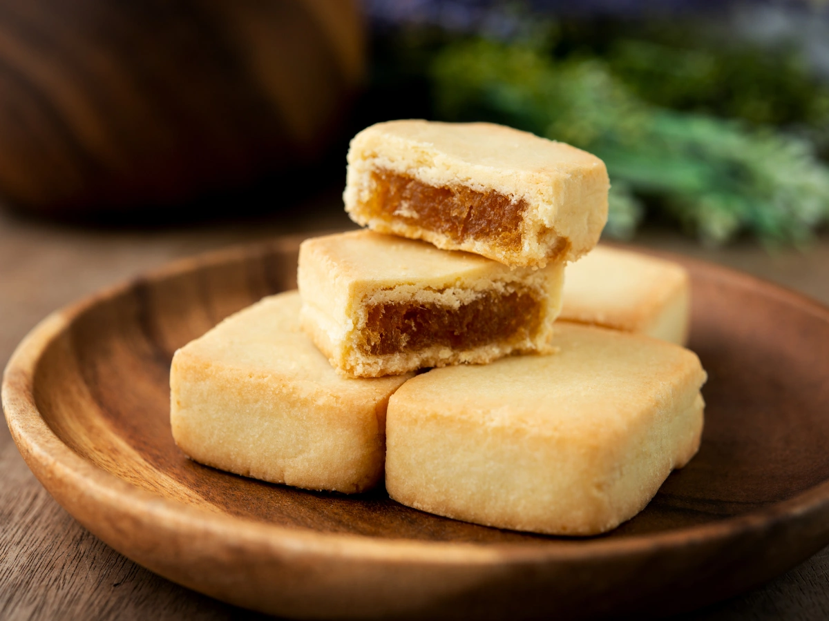 A wooden plate holding four square pastries, one broken open to reveal a fruit paste filling, possibly pineapple, with blurred greenery in the background.