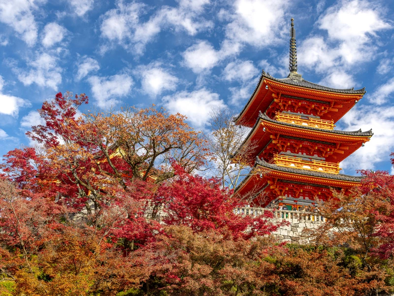 Bright red and orange autumn foliage surrounding the three‑story pagoda at Kiyomizudera Temple in Kyoto, Japan, under a blue sky with scattered clouds.
