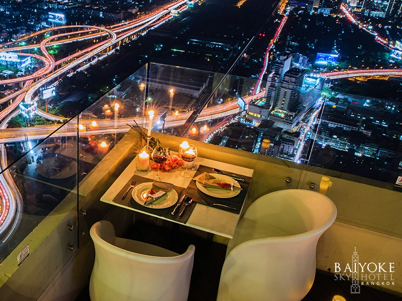 The image shows a nighttime view from a high vantage point, likely from a rooftop or high-rise building. There is a table set for two with plates of food, candles, and utensils. The table is positioned near glass barriers overlooking a cityscape with illuminated roads and buildings. The text "Baiyoke Sky Hotel Bangkok" is visible in the bottom right corner of the image.