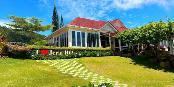 Jerai Hill Resort building with a red-tiled roof and large glass windows, surrounded by manicured gardens and a lush green lawn. A sign reading “I ♥ Jerai Hill” stands at the front under a bright blue sky.