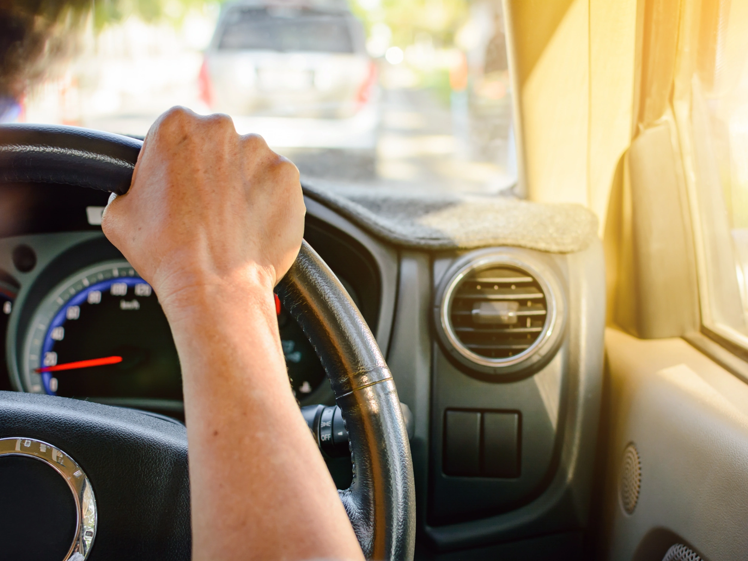 A person driving a car, with the focus on the person's hand gripping the steering wheel. The dashboard and part of the air vent are visible, and there is another vehicle ahead on the road. The scene is well-lit, suggesting it is daytime