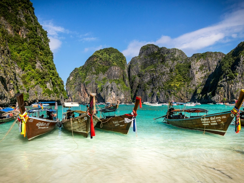 Traditional long-tail boats anchored in clear turquoise water near limestone cliffs covered in greenery under a bright blue sky.