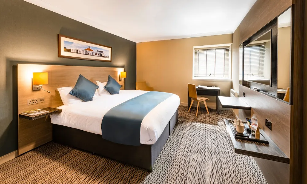 Modern guest room at Copthorne Aberdeen featuring a king-size bed with white linens and blue accents, wall-mounted TV, work desk, and warm contemporary decor