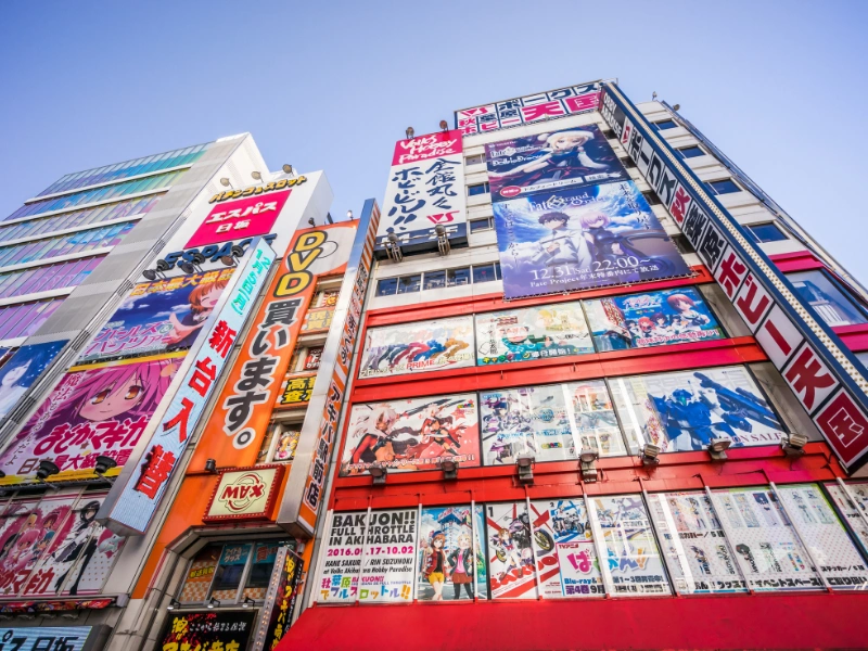 Street view of Akihabara, Japan, with tall buildings covered in colorful advertisements and anime posters promoting DVDs and entertainment products.