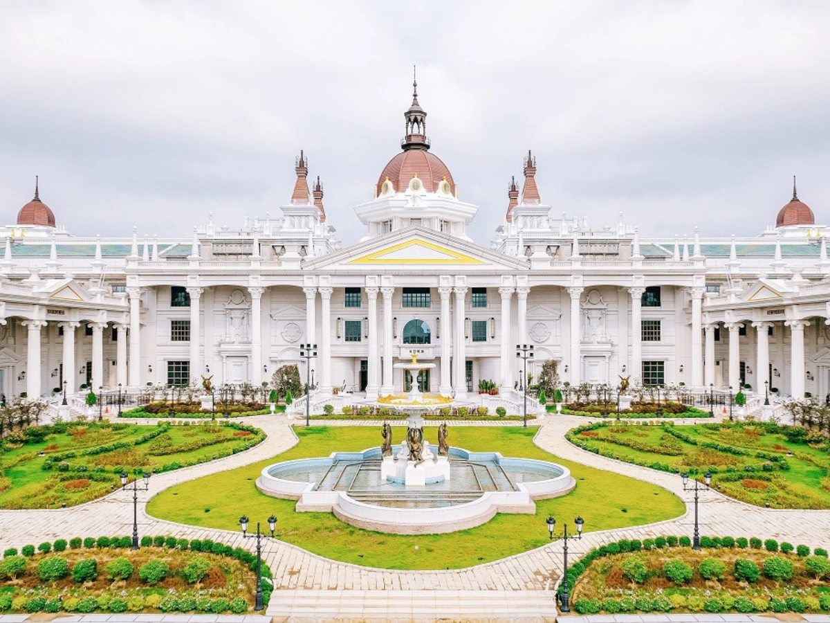 A grand white building with classical architecture, featuring a large central dome, smaller side domes, and multiple columns, set behind a landscaped garden with green lawns, pathways, a central fountain, and statues.