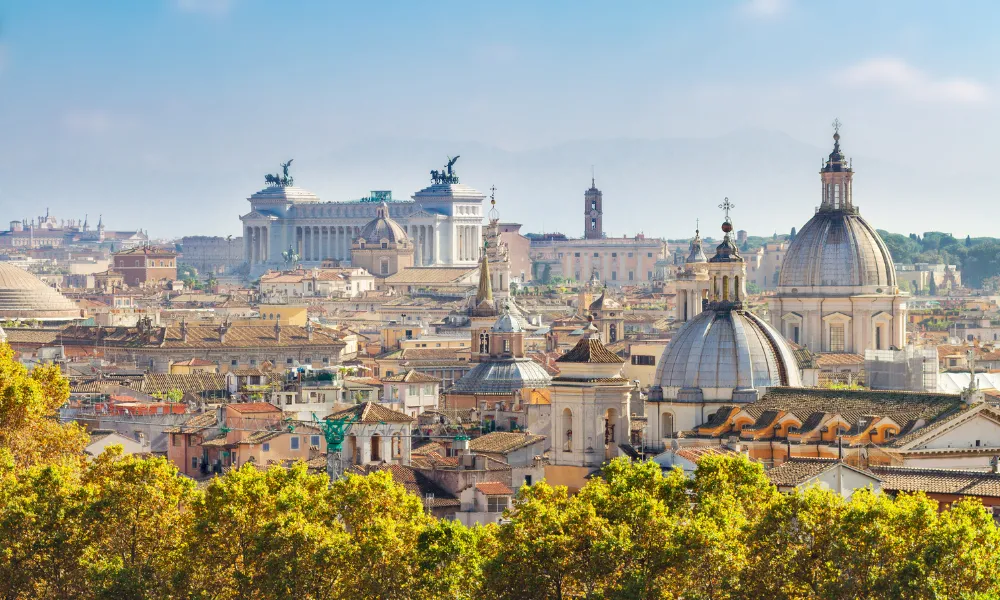 A panoramic view of Rome showcasing a mix of historical and modern architecture. Prominent landmarks include the Altar of the Fatherland with its statues and columns, and several domed churches. The foreground features lush green trees.