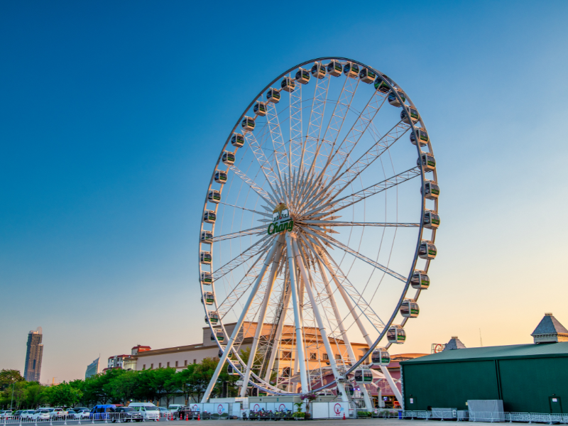 Large Ferris wheel with enclosed cabins set against a clear blue sunset sky, located in an urban area with buildings and trees in the background—fun and safe for children
