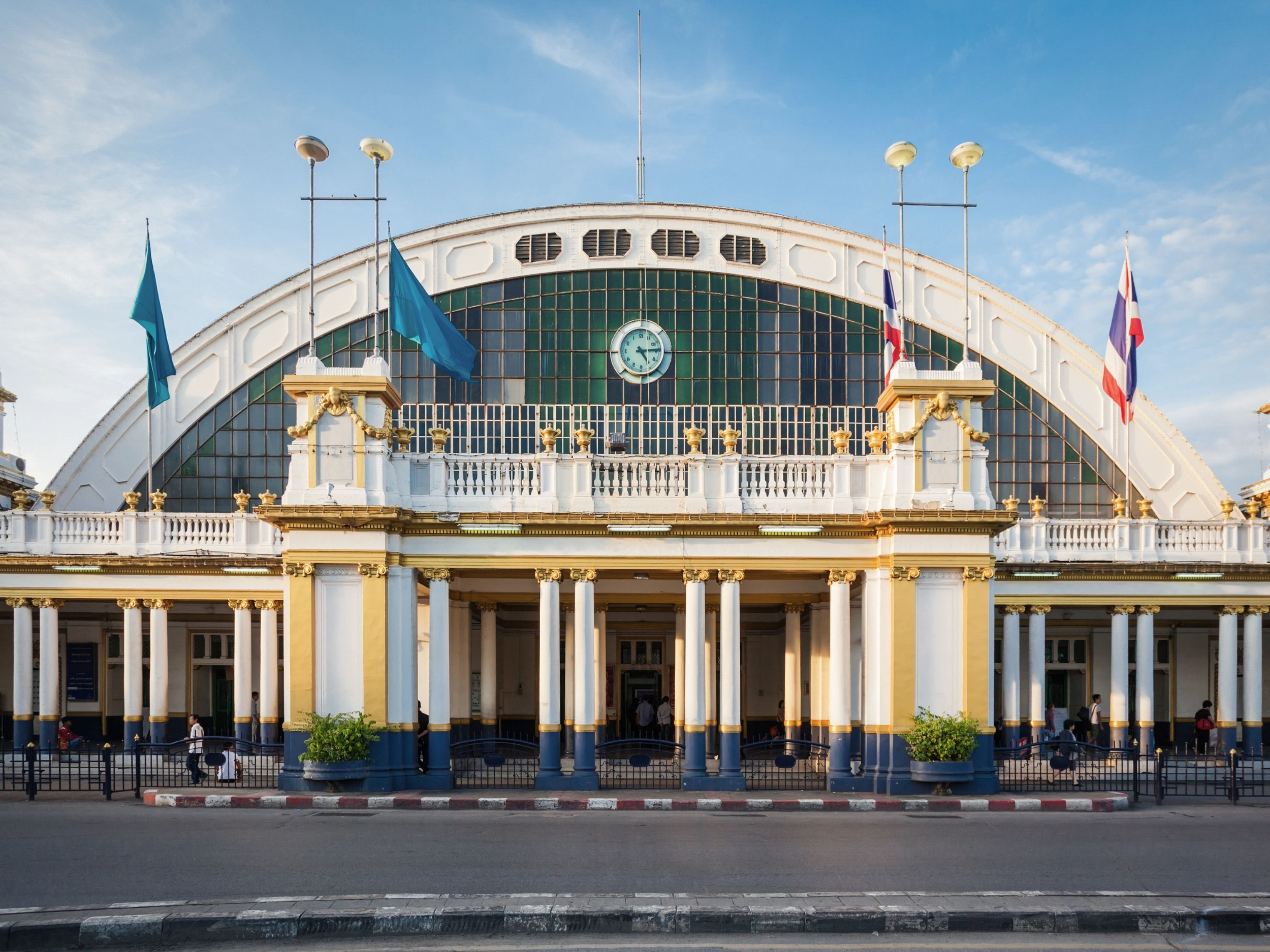 Front facade of Hua Lamphong Railway Station in Bangkok, featuring a large arched window with a central clock, white and gold columns, and flags on poles.