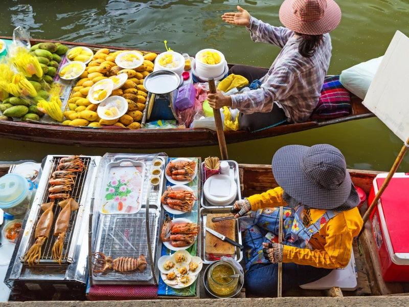 Two people on boats at a floating market. One boat is filled with various fruits, including mangoes and pineapples, while the other boat has seafood dishes such as grilled prawns and squid. The person on the fruit boat is wearing a pink hat and gesturing with one hand, while the person on the seafood boat is wearing a blue hat and preparing food.