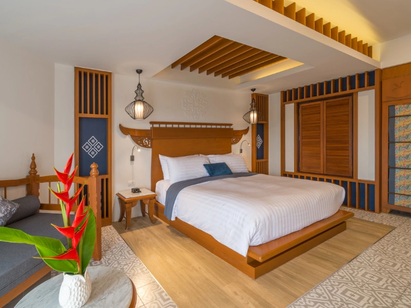 A well-decorated bedroom with a large bed in the center. The bed has white linens, a blue throw blanket, and several pillows. There are two hanging lamps on either side of the bed, and wooden slats on the ceiling above it. To the left of the bed is a small wooden nightstand with a phone on it. There is also a seating area with cushions to the left of the nightstand. In front of this seating area is a round table with red flowers in a white vase. The room features intricate woodwork and patterns on the walls and floor, giving it an elegant and cozy atmosphere.