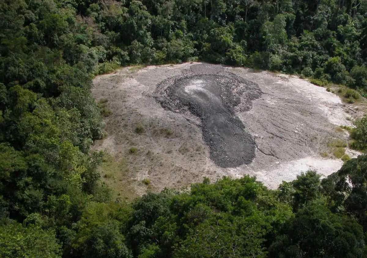 A clearing in the forest featuring the Lipad Mud Volcano, with soft grey mineral-rich mud spread across the ground, surrounded by sparse vegetation and tropical trees.