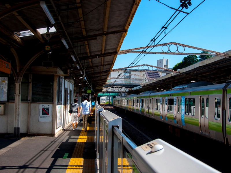 Train station platform with a train on the right, two people walking toward the end, yellow tactile paving along the edge, and overhead wires above the tracks.