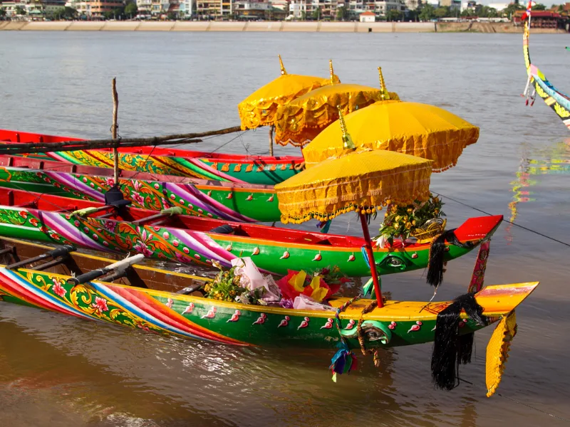 Colorful decorated boats docked on a river, each adorned with vibrant patterns, yellow parasols, and flowers, with buildings visible along the riverbank in the background.