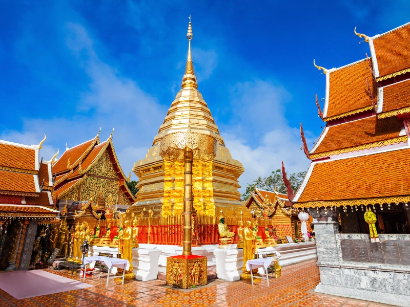 Buddhist temple complex featuring a central golden stupa, traditional Thai architecture with tiered roofs, Buddha statues, and offerings, set against a bright blue sky.