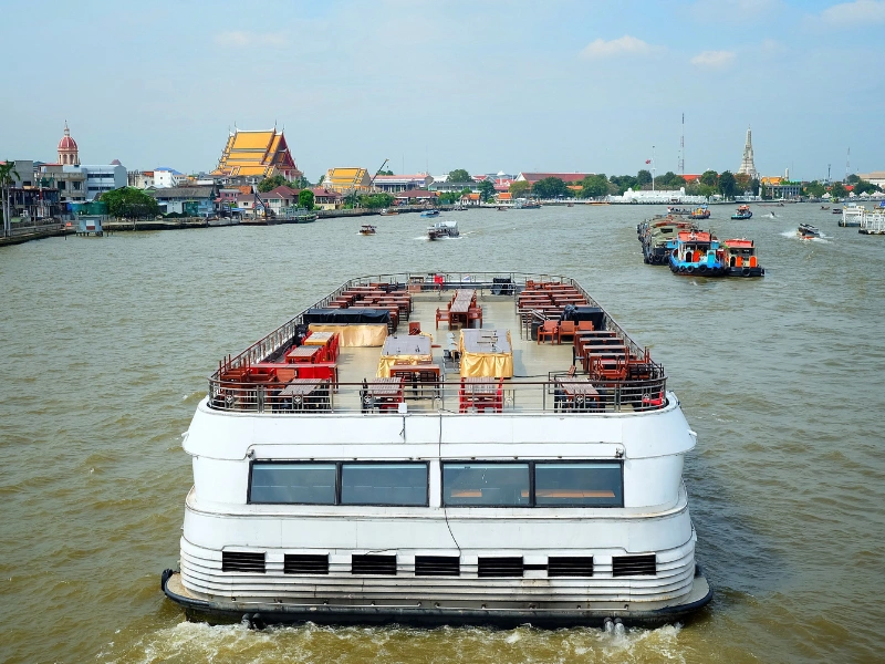 The image shows a large white boat or ferry on a river, viewed from behind. The boat has an open deck with seating and tables arranged in rows. In the background, there are several other boats on the river, and along the riverbank, there are various buildings including temples with distinct architectural features. The sky is clear with some clouds visible.