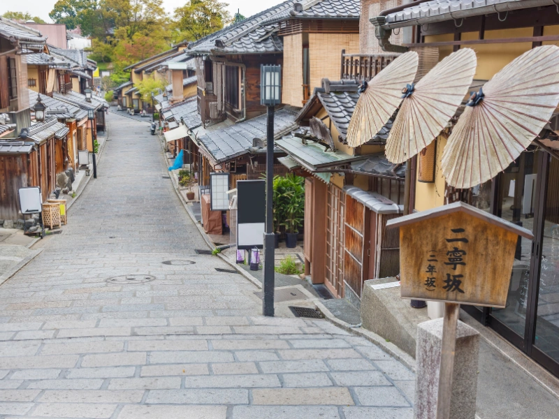 Quiet traditional street in Kyoto, Japan, lined with wooden buildings and tiled roofs, featuring paper umbrellas and a wooden signpost labeled ‘Ninenzaka’.