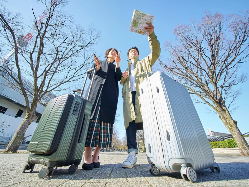 Two people standing outdoors with a green and silver suitcase, looking at a map or guidebook under a clear blue sky with leafless trees in the background.