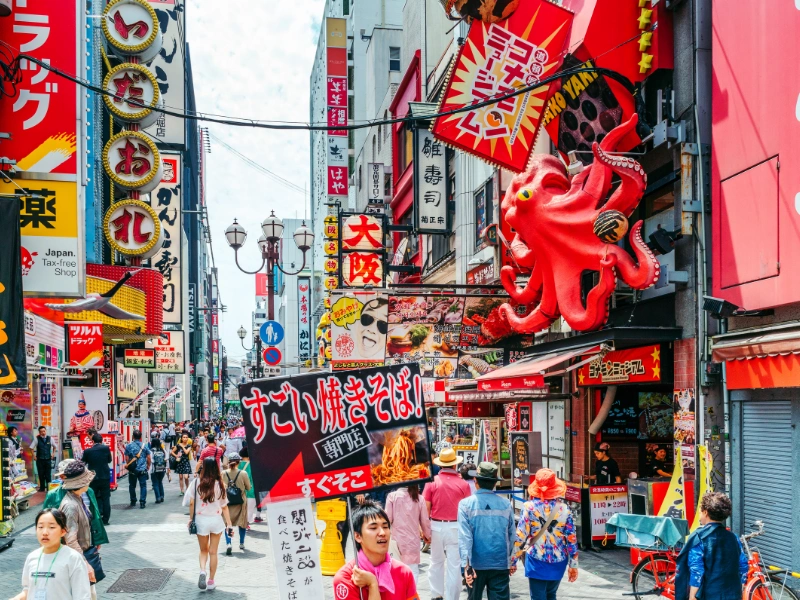 Lively street scene in Dotonbori, Osaka, with colorful Japanese signs including a large red octopus, and crowds of people walking among shops and eateries.