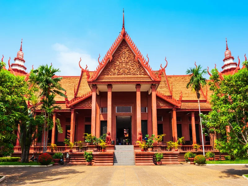 A traditional Southeast Asian temple with a red-tiled roof, ornate carvings, and spires, supported by multiple columns and surrounded by lush greenery and potted plants.