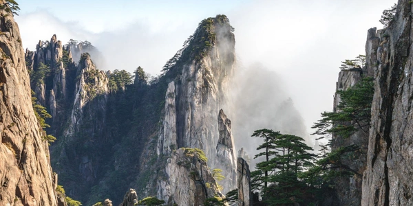 A panoramic view of Huangshan Mountain in eastern China, featuring rugged granite formations rising above drifting mist and surrounded by resilient pine trees. The UNESCO World Heritage Site is celebrated for its stunning natural scenery, hiking trails, and eco-tourism experiences.