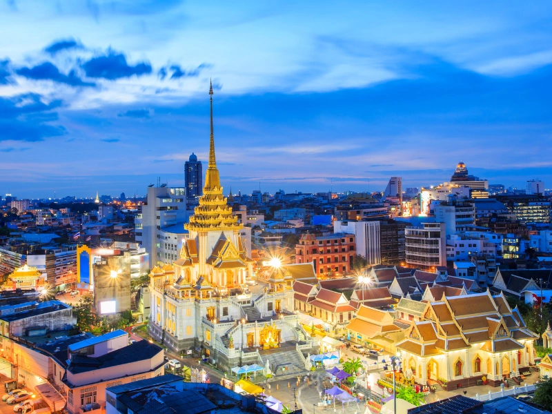 Aerial view of a cityscape at twilight, centered around a golden temple with a tall spire, surrounded by illuminated buildings under a partly cloudy sky with hues of blue and orange.