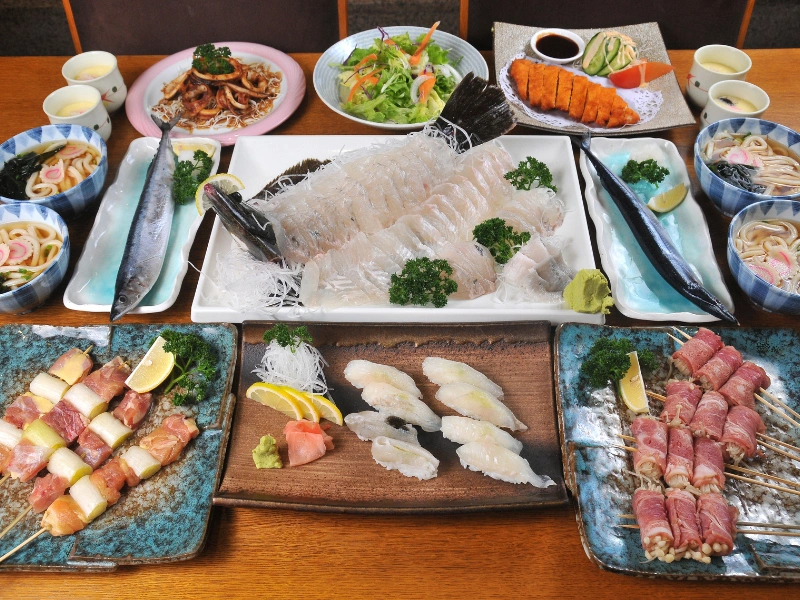 A beautifully arranged kaiseki meal featuring a central platter of sashimi, surrounded by sushi, skewered meats, salads, soups, and dipping sauces, all garnished with lemon wedges and parsley.