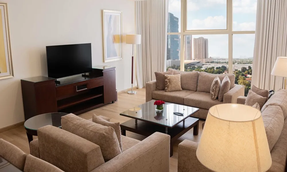 Modern living room at Grand Millennium Dubai featuring beige sofas, a flat-screen TV, large windows with city views, and elegant décor with natural light.