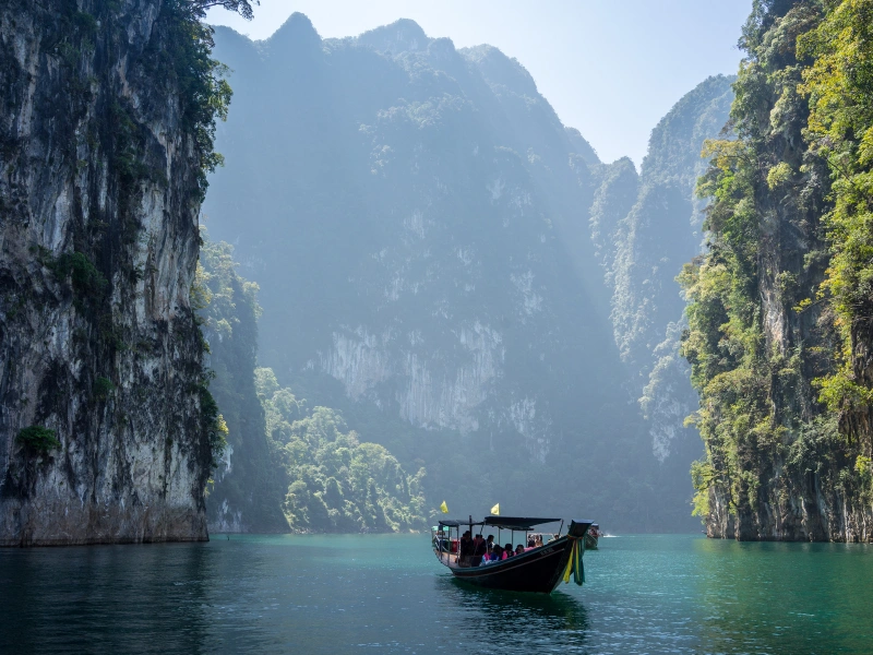 A boat with people floats on calm water between steep, lush green cliffs under a clear blue sky.