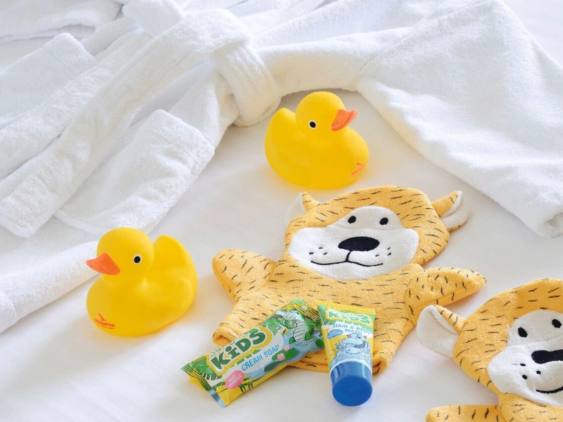 Three yellow rubber ducks, a white bathrobe, a tiger-shaped stuffed toy, and two tubes of toothpaste labeled 'KIDS' arranged on a white surface.
