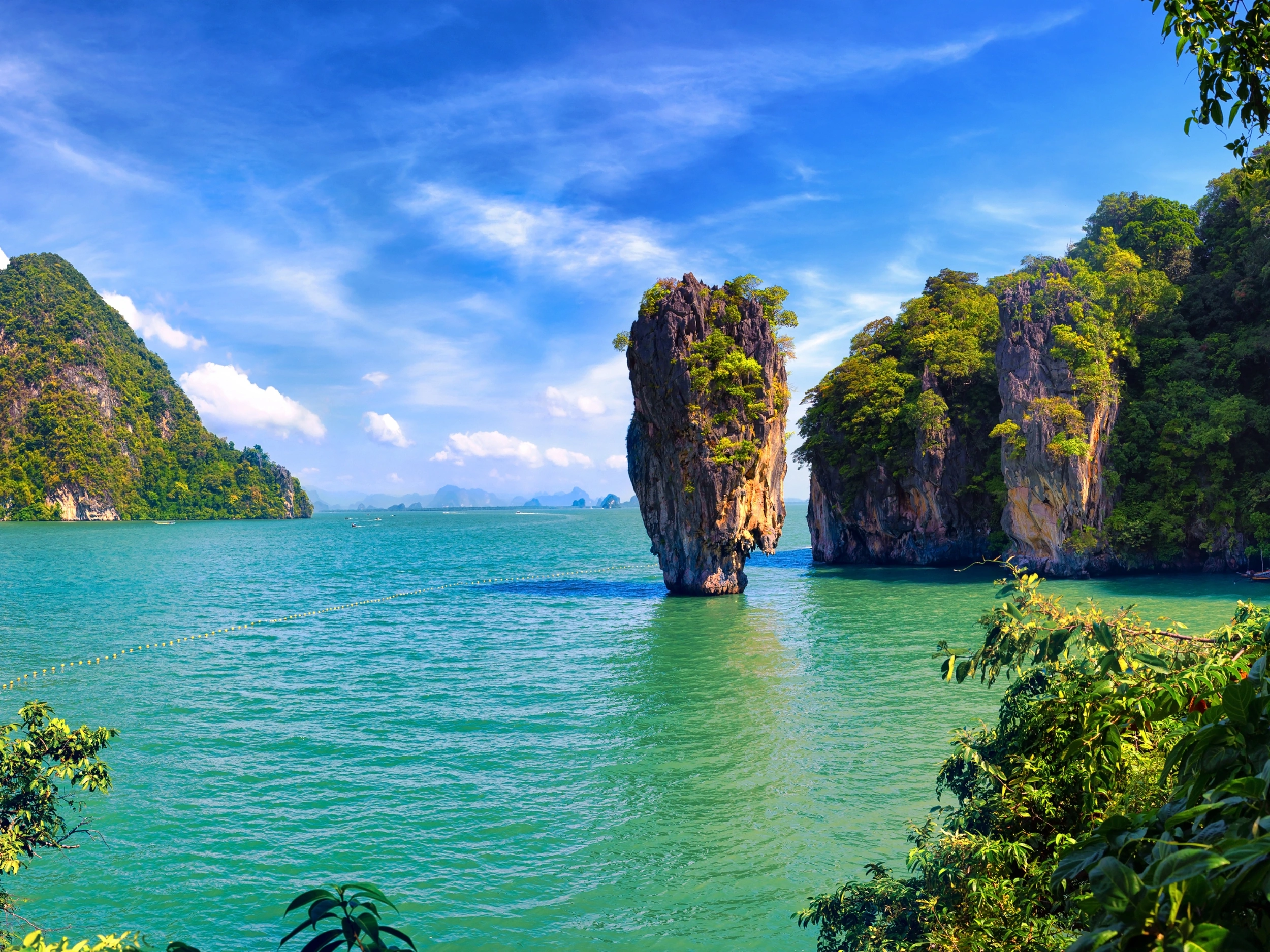 The image shows a scenic view of a tropical seascape with clear blue skies and turquoise waters. There are two prominent limestone karst formations rising from the water, covered with lush green vegetation. In the background, there are more islands and rock formations visible in the distance. The foreground includes some greenery and foliage along the edge of the water.