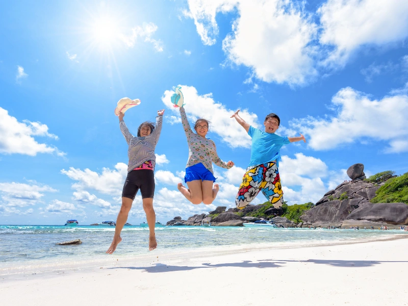 The image shows three people jumping in the air on a sandy beach with clear blue skies and scattered clouds. The ocean is visible in the background, along with some rocks and greenery on the right side of the image. The sun is shining brightly, creating a vibrant and cheerful atmosphere.
