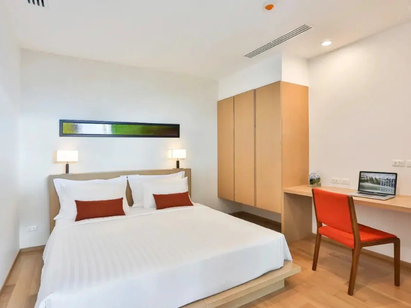 Modern minimalist hotel bedroom featuring a large bed with white bedding and red accent pillows, wall-mounted lamps, green and black artwork above the bed, a wooden desk with an orange chair and open laptop, light wood flooring, and a built-in wardrobe.