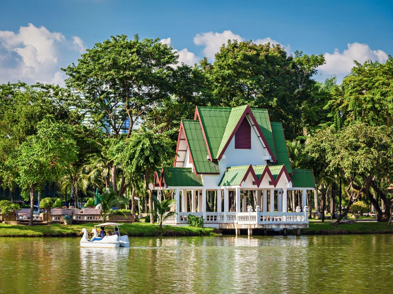 Scenic lakeside house with green roofs and white walls, red-trimmed gables, surrounded by lush trees, with a paddle boat carrying two people on the lake under a partly cloudy blue sky.