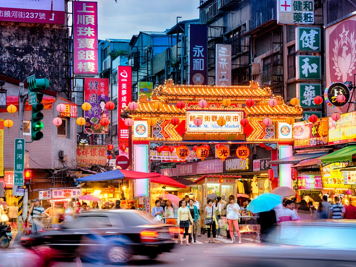 A lively night market scene in an East Asian city, featuring a brightly lit entrance with traditional architectural elements, hanging lanterns, Chinese signage, bustling crowds, and various stalls and shops along the street.