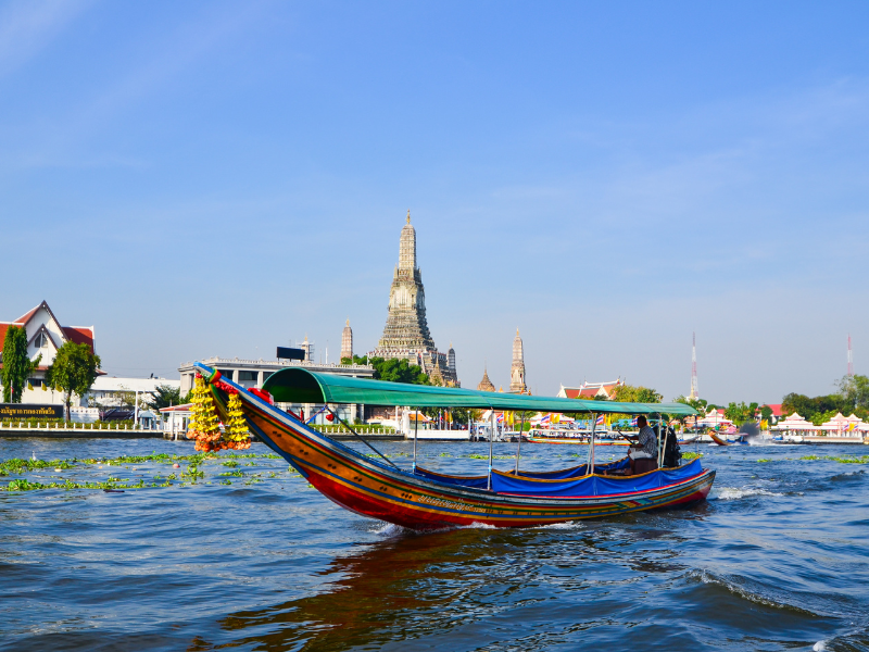 olorful long-tail boat on a river with Wat Arun temple in the background under a clear blue sky, surrounded by buildings and greenery.