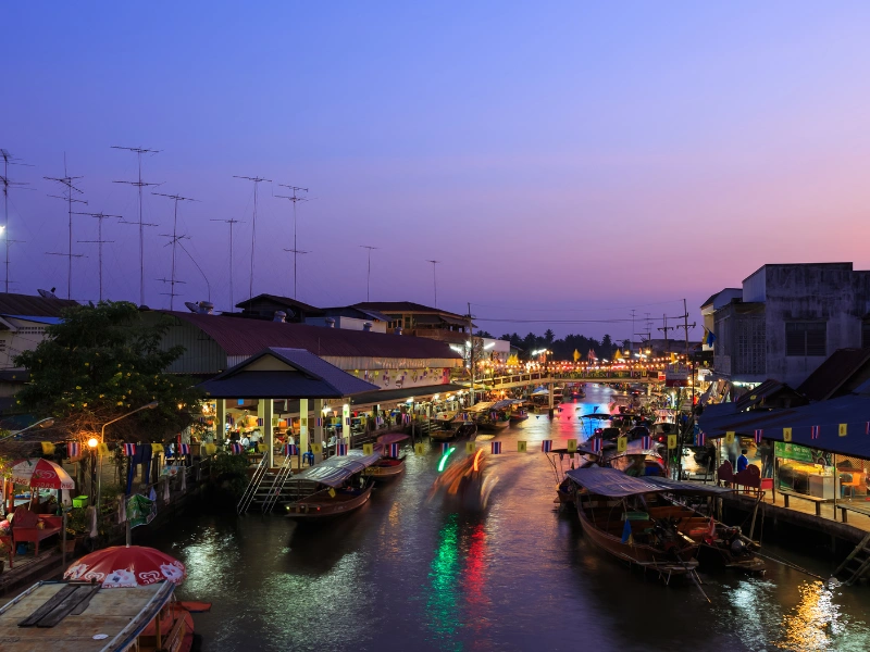 A vibrant evening scene of a floating market. There are numerous boats on the water, some with canopies, and many people are visible on the boats and along the waterfront. The market is bustling with activity, with various stalls and shops lining both sides of the canal. The sky is transitioning from dusk to night, displaying shades of blue and purple. Bright lights from the market reflect on the water, adding to the lively atmosphere.