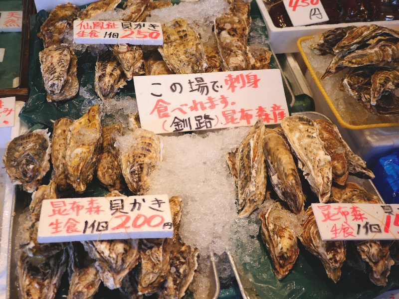 Seafood stall at Nijo Market with shellfish on ice and handwritten Japanese price signs.
