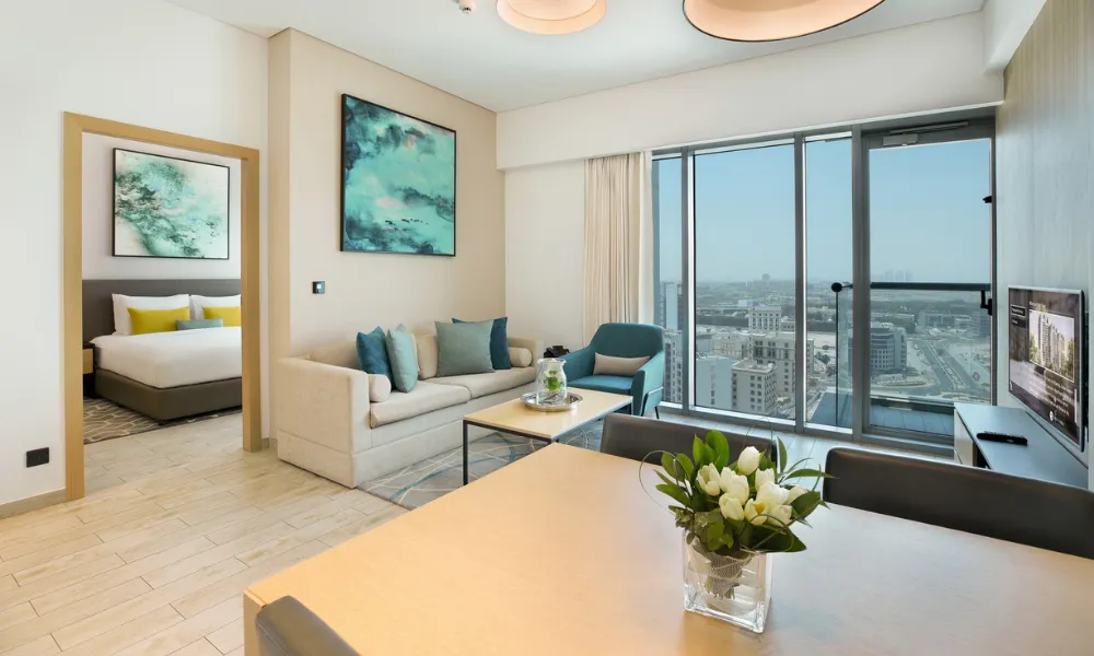 Modern serviced apartment at Millennium Montrose Dubai featuring a bright living area with sofa and armchair, dining table with floral centerpiece, large windows with city views, and a separate bedroom.