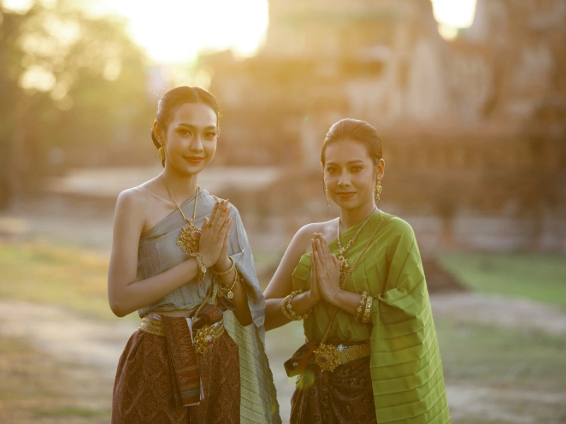 Two individuals in traditional Thai attire standing side by side with hands together in a greeting gesture, outdoors during sunset with blurred background elements.