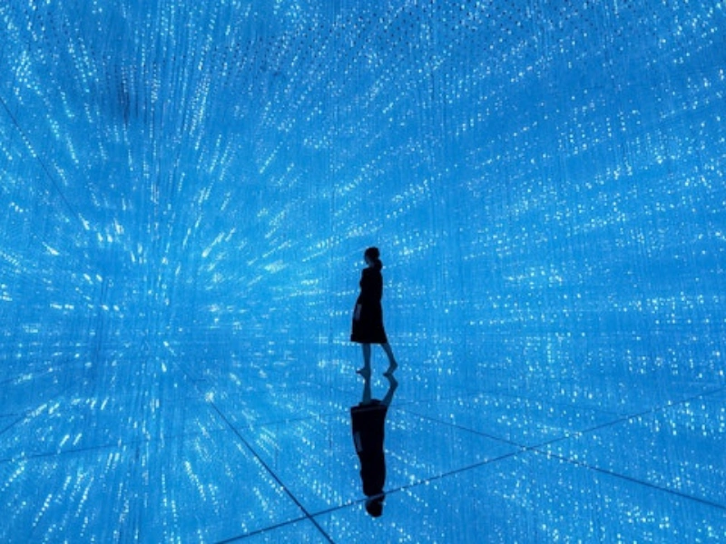Silhouette of a person standing in an immersive room filled with blue grid-pattern lights on all surfaces, creating a starry, infinite space effect with reflections on the shiny floor.