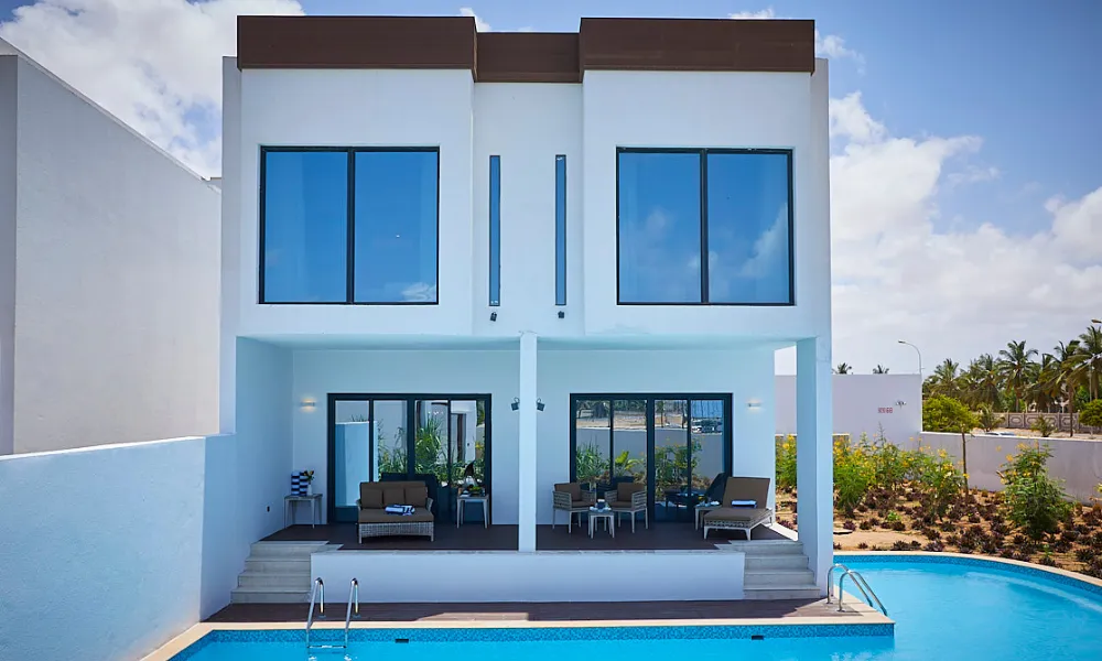 Modern white villa at Millennium Salalah Oman featuring two floors, large glass windows, private terrace with outdoor seating, and a swimming pool under clear blue skies.