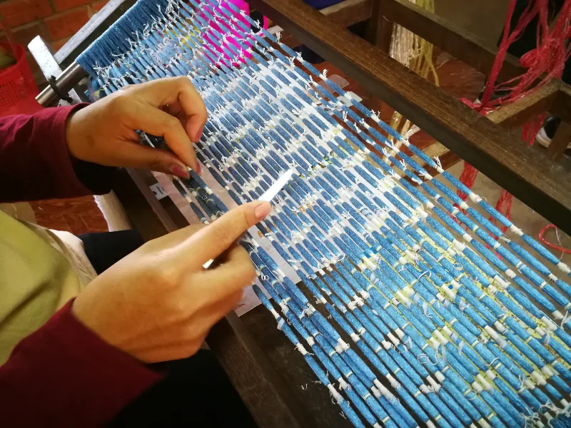 A person weaving fabric on a wooden loom, manipulating blue, white, and yellow threads with their hands, surrounded by various threads in the background.